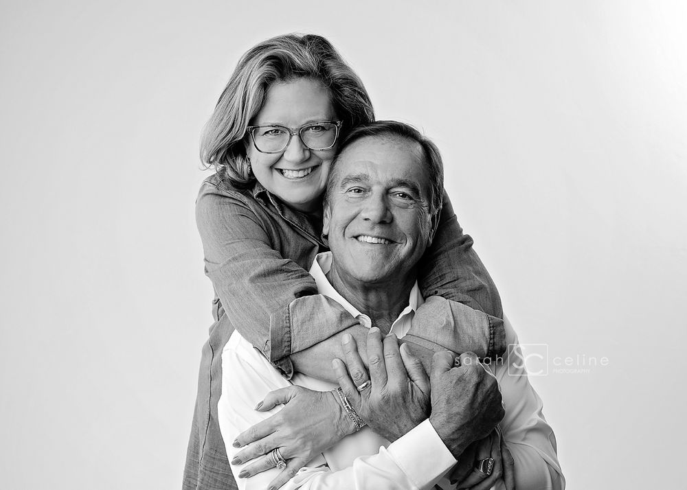 Black and white photo of a smiling couple, woman hugging man from behind, both wearing glasses, studio portrait.