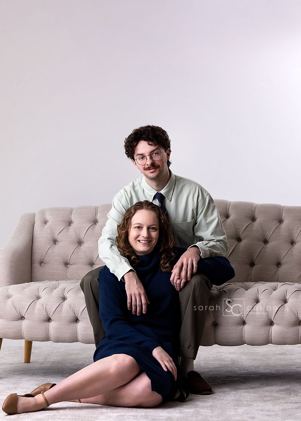 Couple in vintage-inspired clothing pose together on a tufted beige couch in a minimalist setting.
