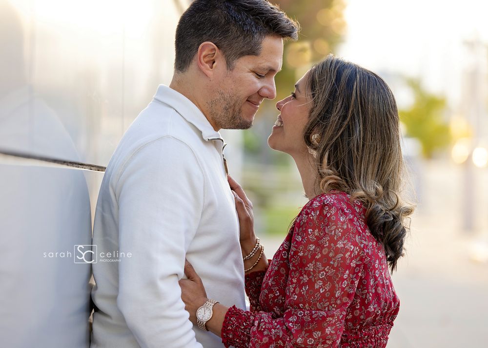 Smiling couple embracing outdoors during an engagement photo session, with a soft blurred background and warm golden sunlight.