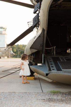 A small child in a white dress explores a military helicopter on a tarmac.