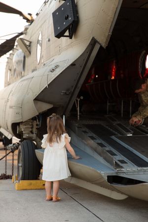 Small child explores military helicopter ramp while wearing white dress and boots.