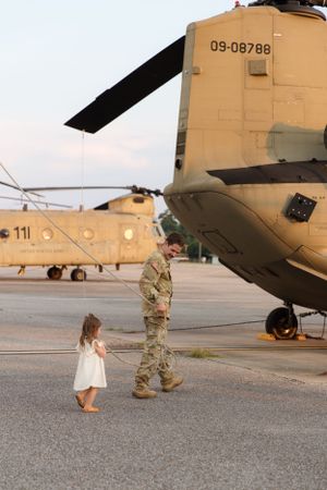 Tan military helicopter on tarmac with figures walking near its side.