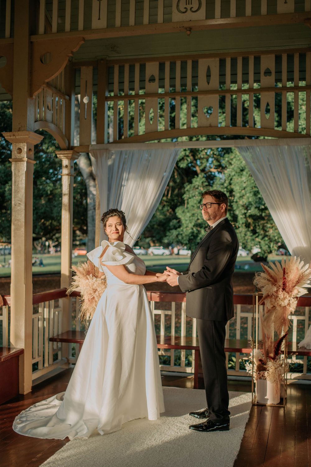 A romantic wedding ceremony takes place in a vintage gazebo decorated with flowing white curtains and pampas grass.