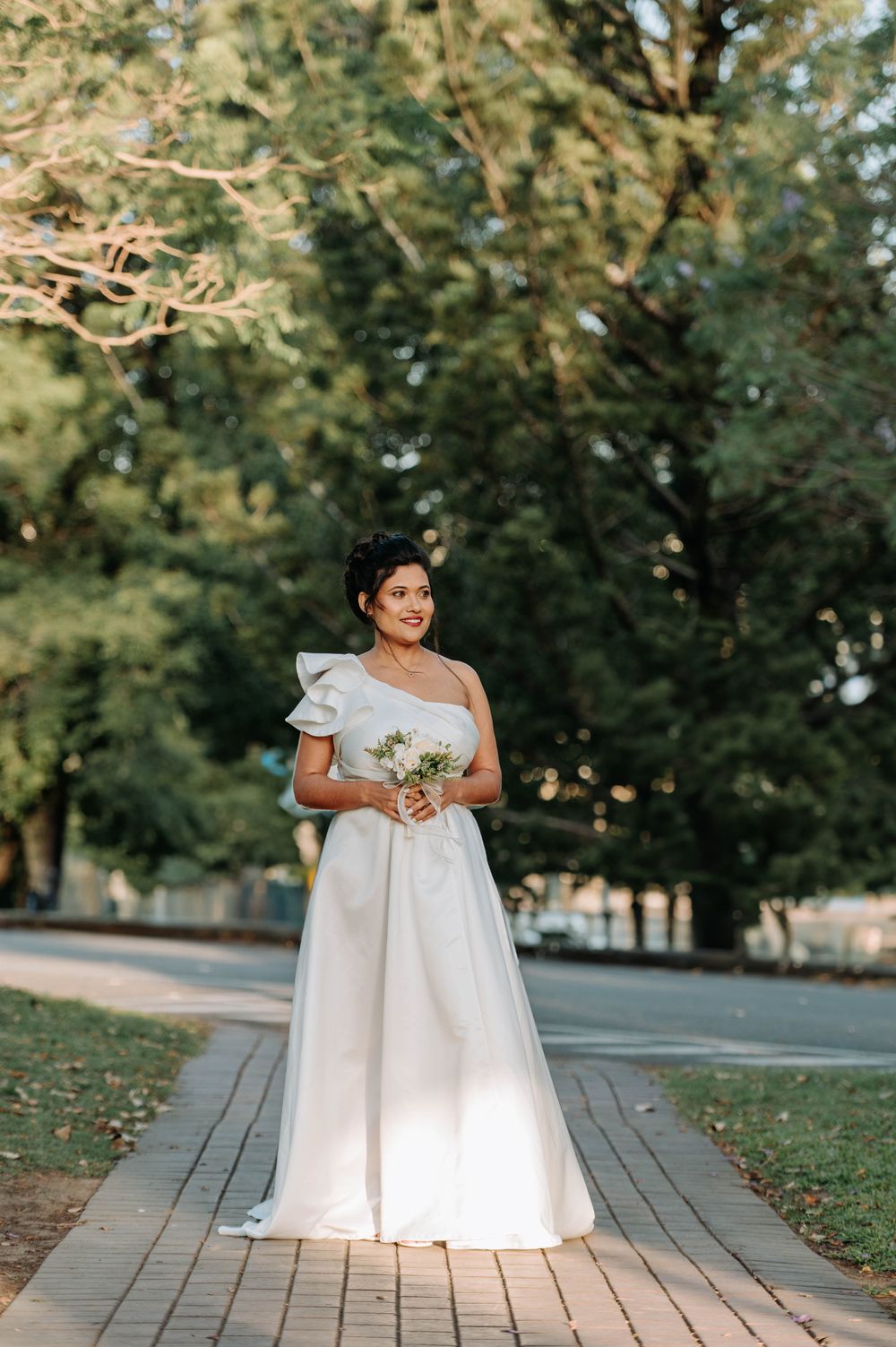 A bride in a one-shoulder wedding gown holds a bouquet while standing on a stone pathway.