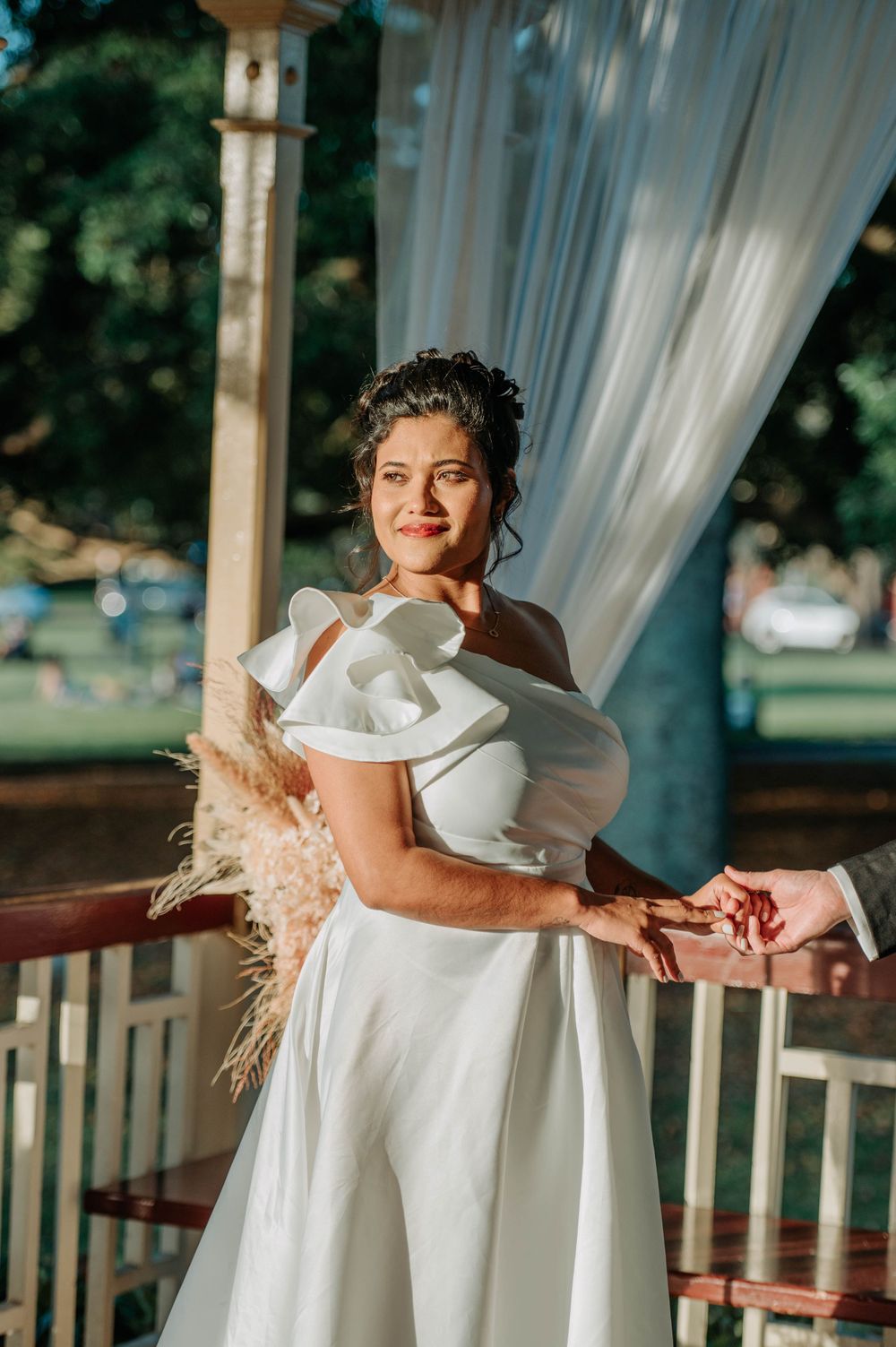 A bride in an elegant one-shoulder white gown poses in a sunlit gazebo with pampas grass decor.