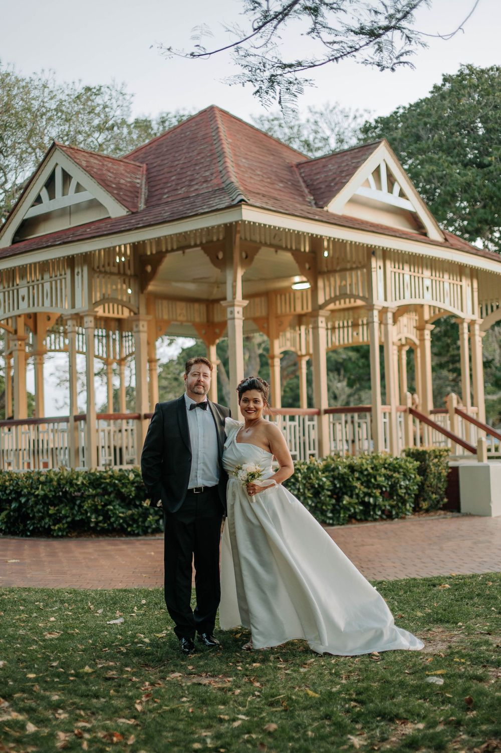 Elegant couple poses in front of a vintage gazebo with ornate wooden details and Victorian-style architecture.