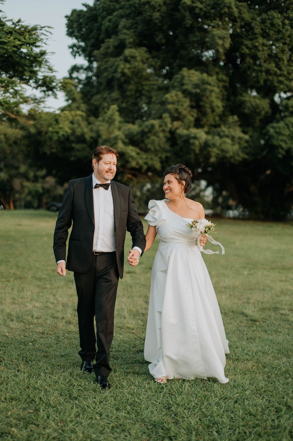 Wedding couple walks hand in hand on lush grass, sharing a joyful moment in their formal attire.