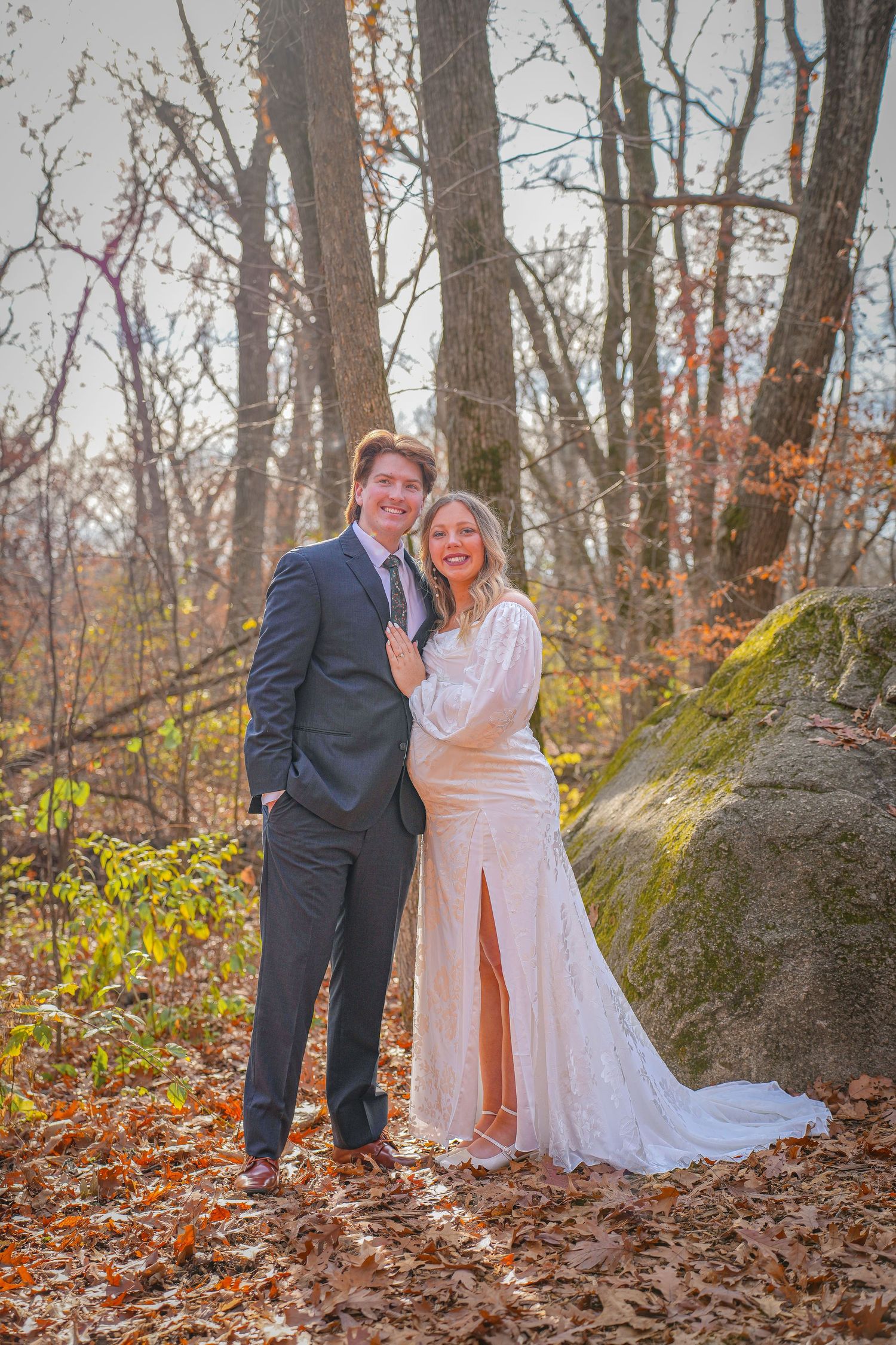 Newlyweds pose together against mossy rock in autumn woodland setting.