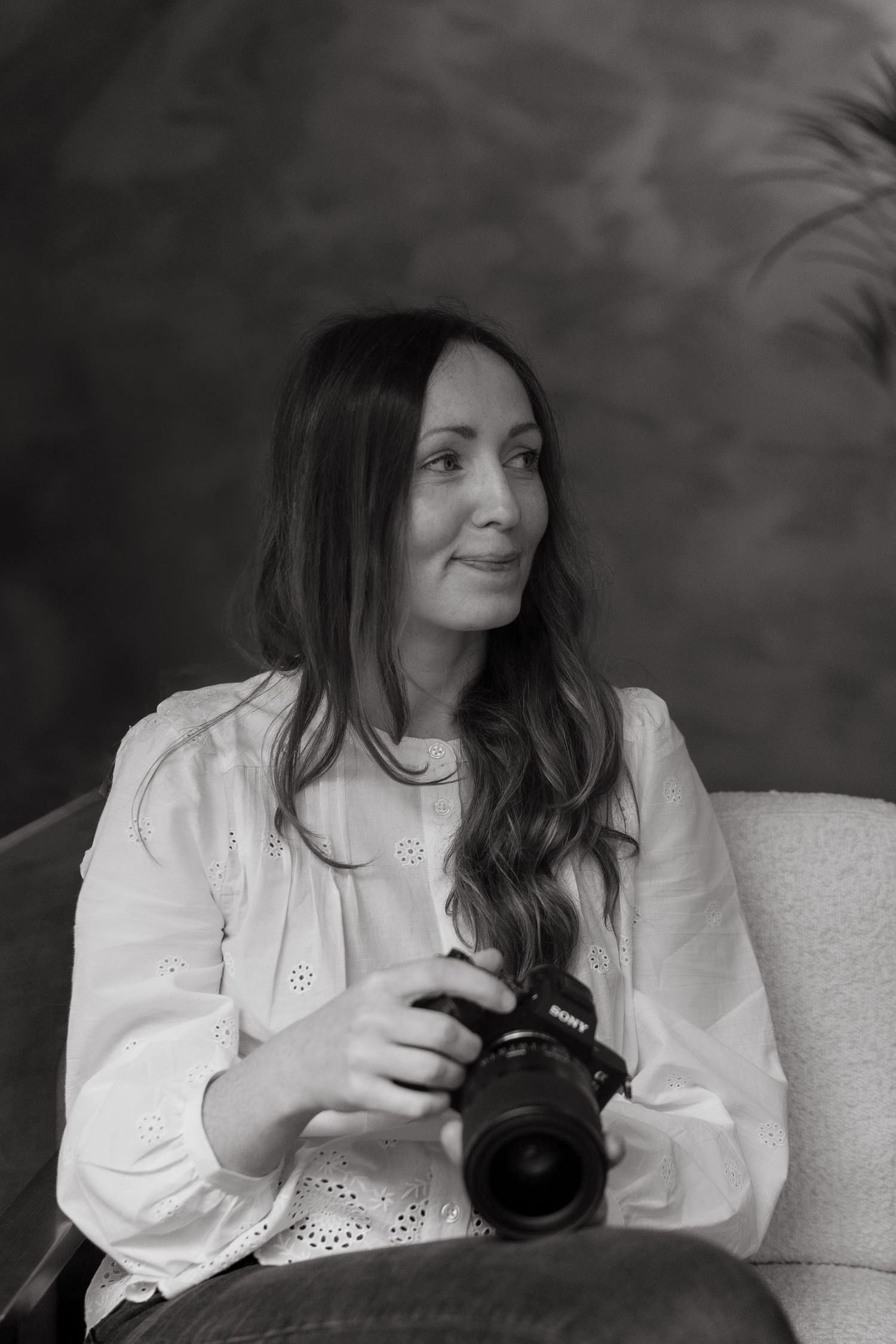 A photographer in a white blouse sits comfortably in an armchair holding a professional camera in warm lighting.