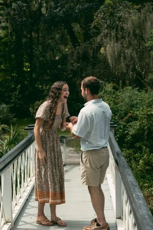 A couple sharing a joyful moment during a proposal on a white bridge surrounded by trees.