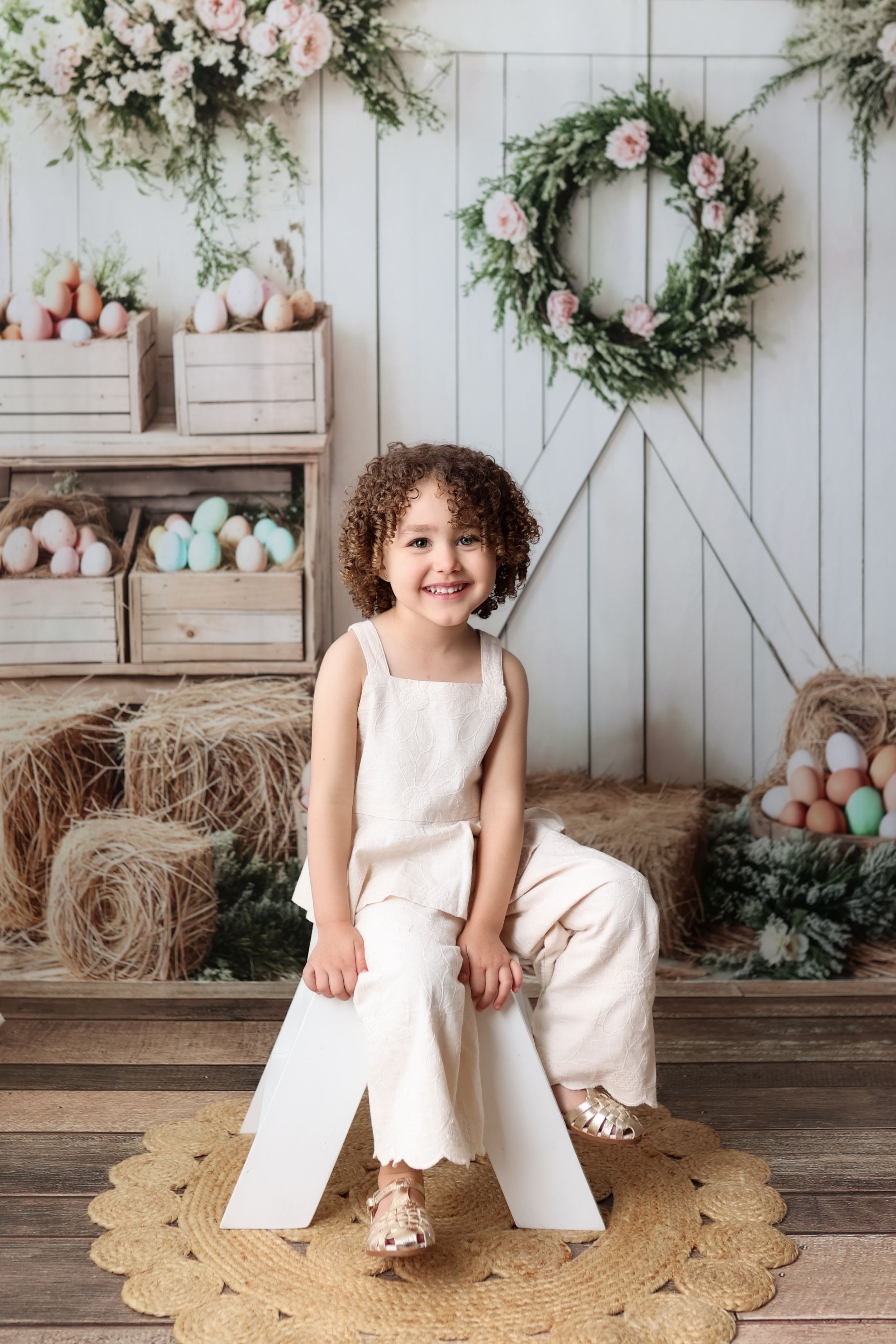 Easter photo backdrop with floral wreath, pastel eggs, hay bales and rustic decor in spring setting.