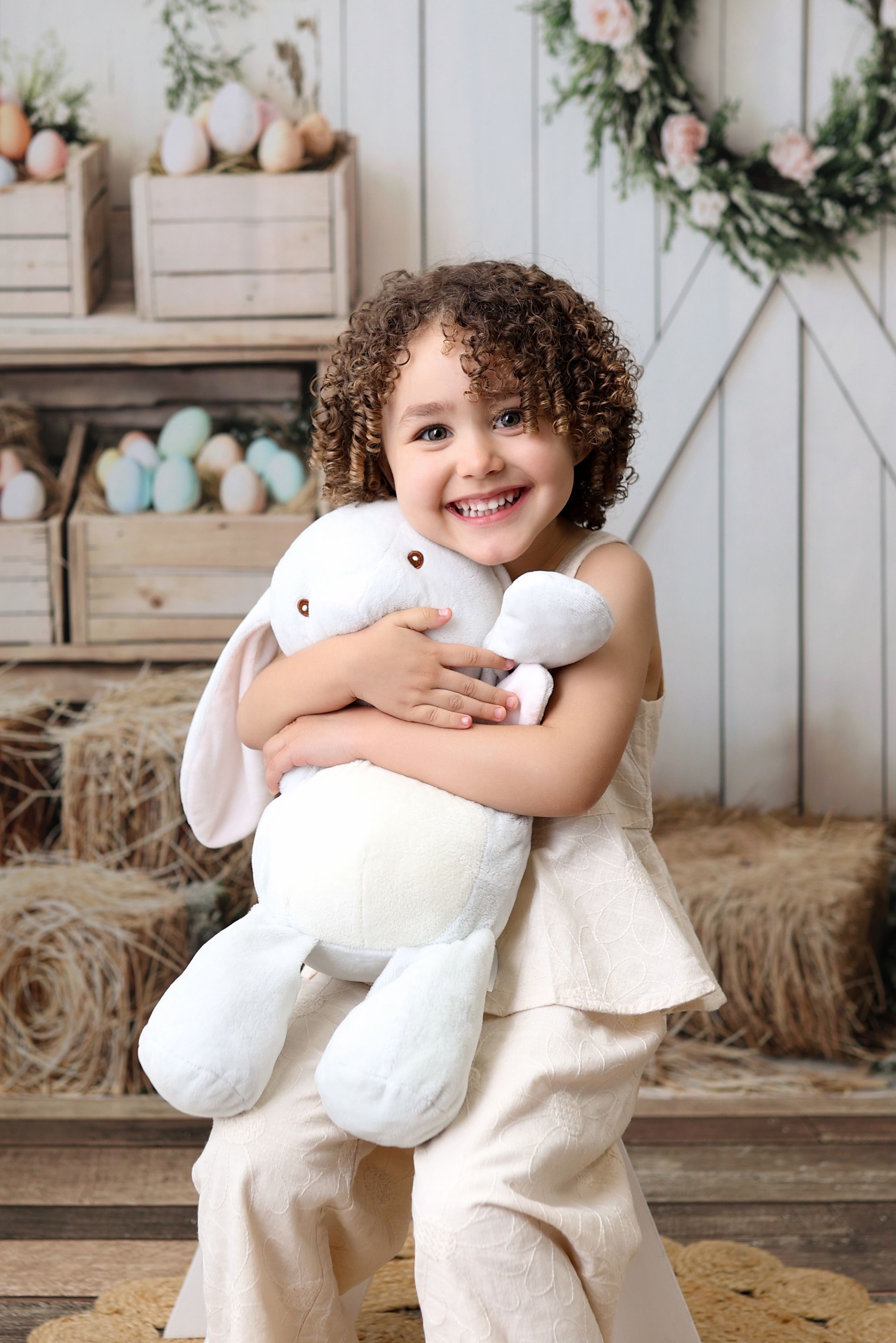 Child in cream outfit hugging large white plush bunny in Easter setting with decorated eggs and floral wreath decor.