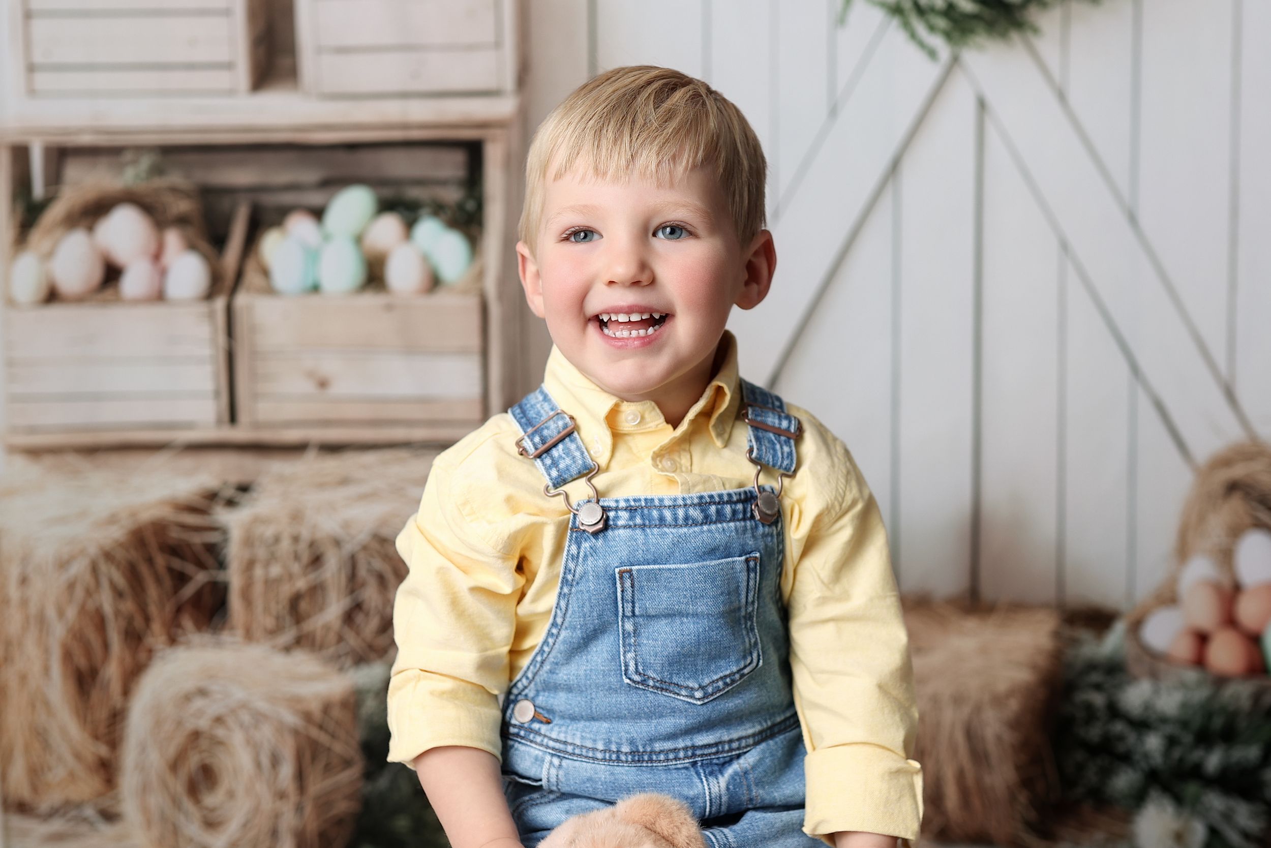Happy child wearing denim overalls and yellow shirt sits on rustic backdrop with hay bales and farmhouse decor.