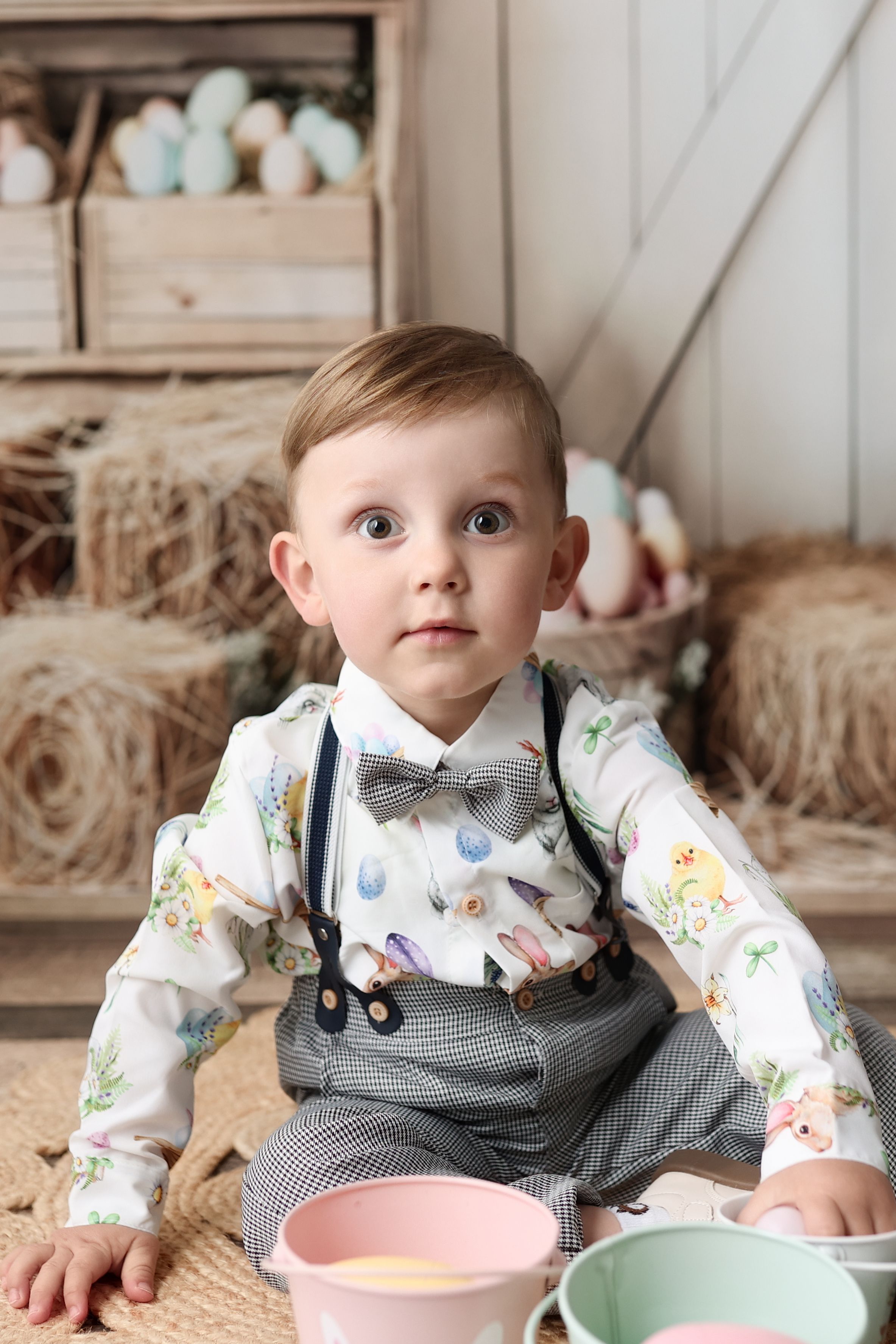 Sweet photo of a child wearing a bow tie and patterned shirt with suspenders sitting by decorative bowls and hay bales.