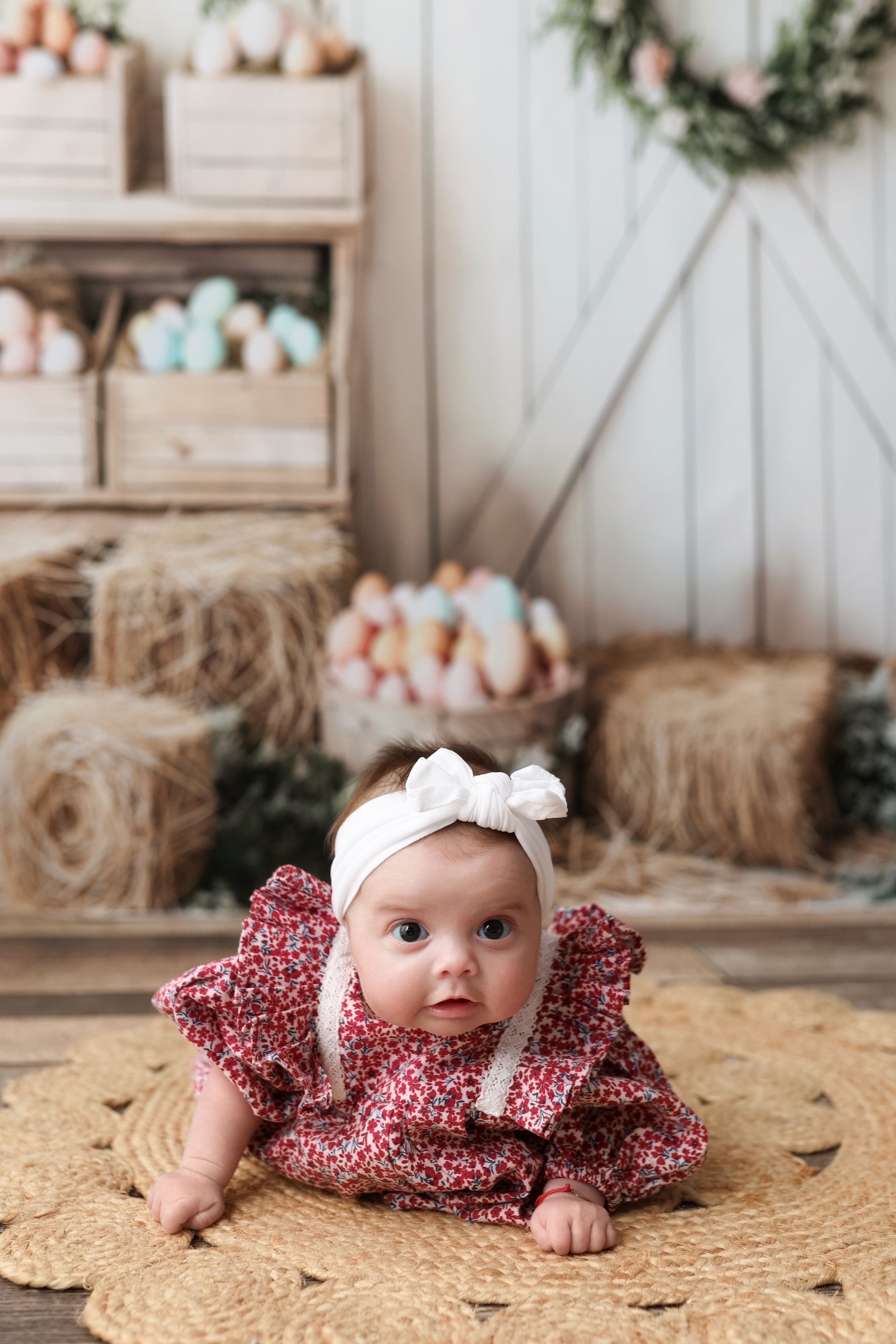 Baby wearing floral dress and white bow headband laying on woven rug in rustic farmhouse setting with hay bales and wreaths.