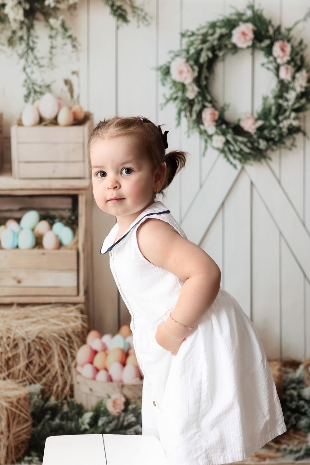 A white rustic spring scene with pastel eggs, a floral wreath, and Easter decor against a white wooden barn door backdrop.
