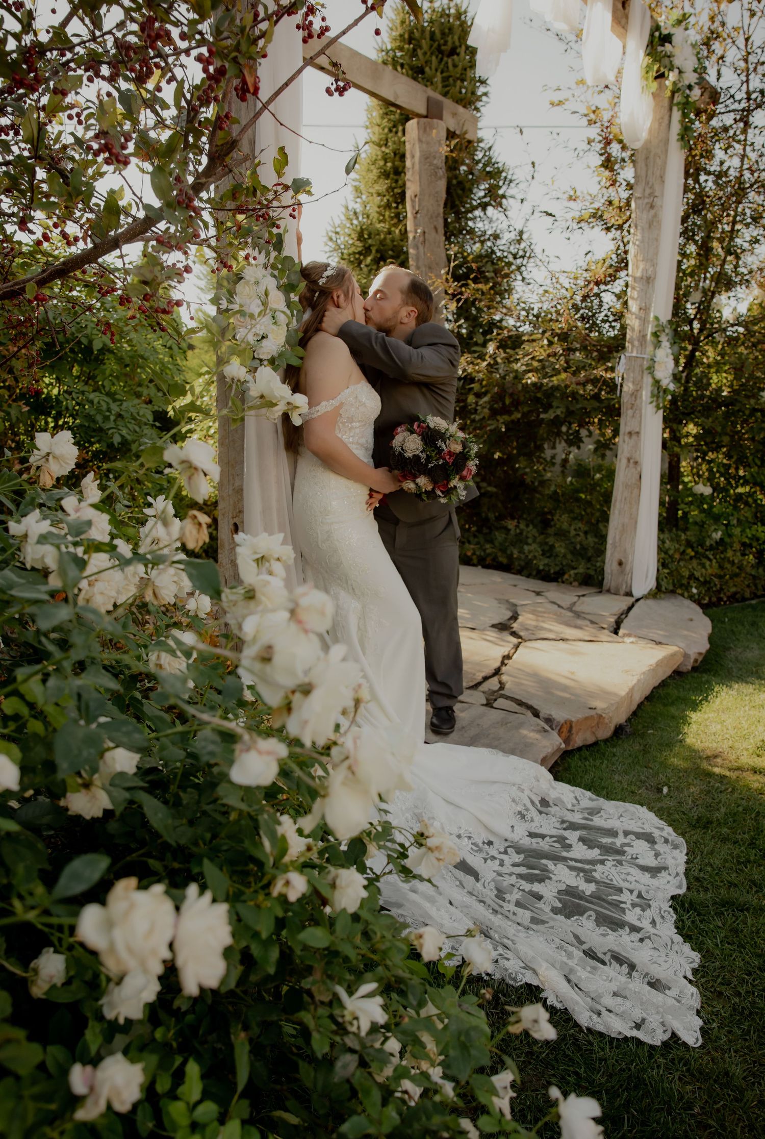 A wedding couple embraces under a flower-adorned arch in a garden setting.
