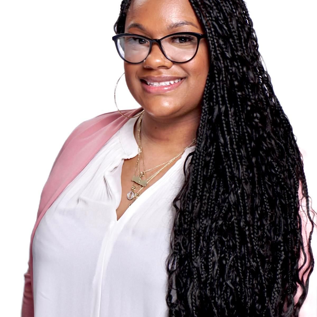 Professional headshot of someone wearing glasses with long braided hair and a white blouse with pink blazer.