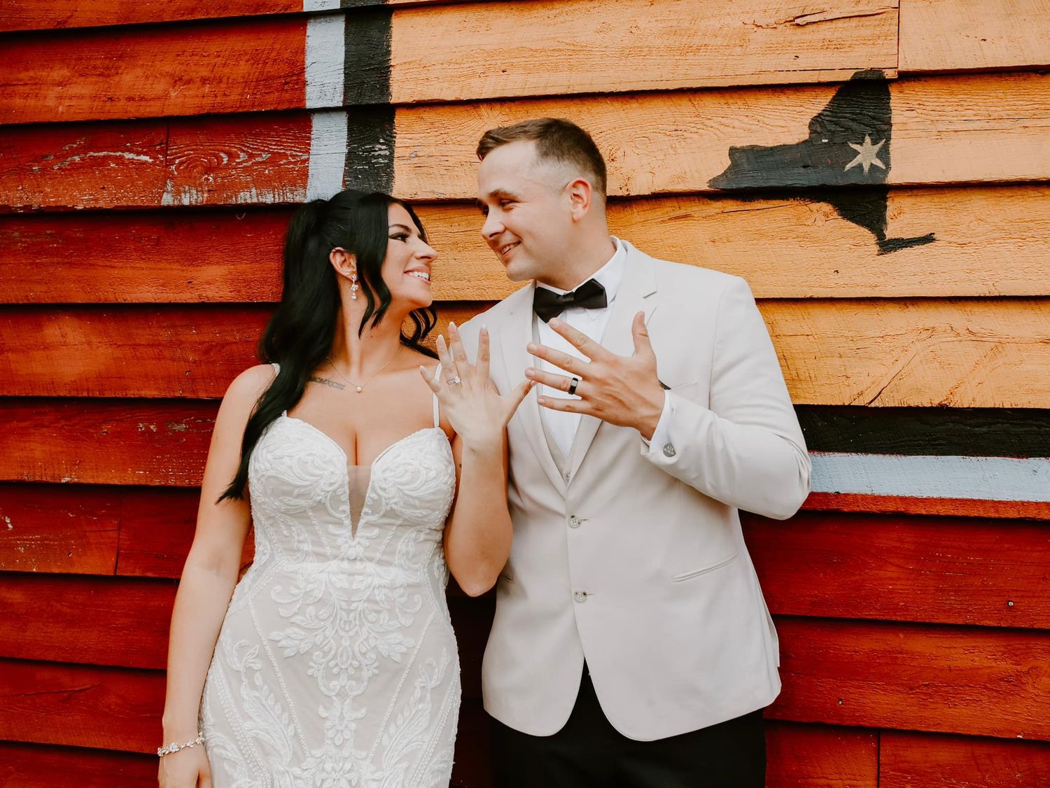 A couple in formal wedding attire standing against a rustic red wood wall in romantic pose.