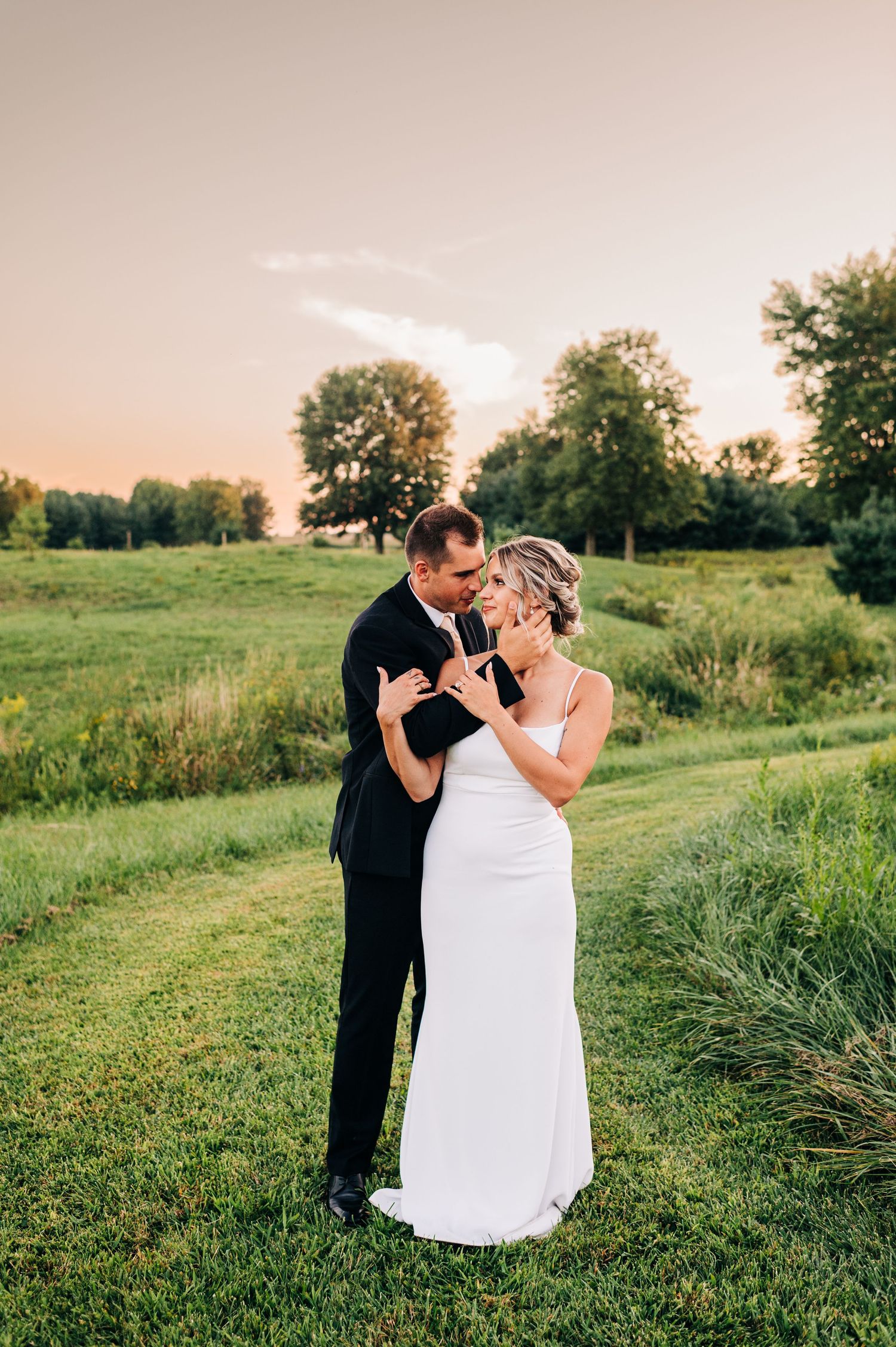 A loving couple shares an intimate moment while dancing together on a grassy field at sunset during their wedding.