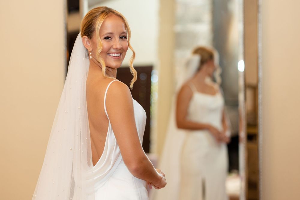 A bride in a backless white wedding dress smiles while looking over her shoulder with a mirror reflection.
