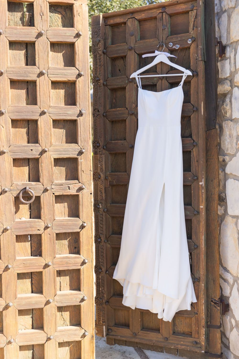 A white long wedding dress hangs on a wooden hanger against a rustic wooden door with geometric patterns.