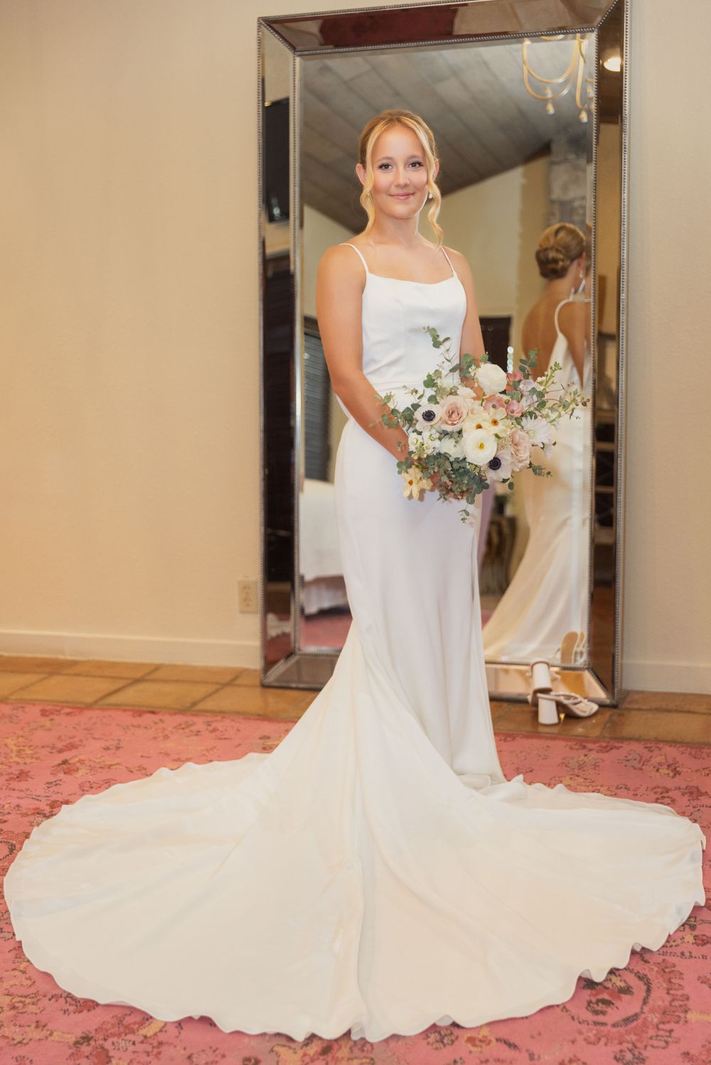 A bride in an elegant white satin gown with long train poses in front of a mirror while holding a pastel floral bouquet.
