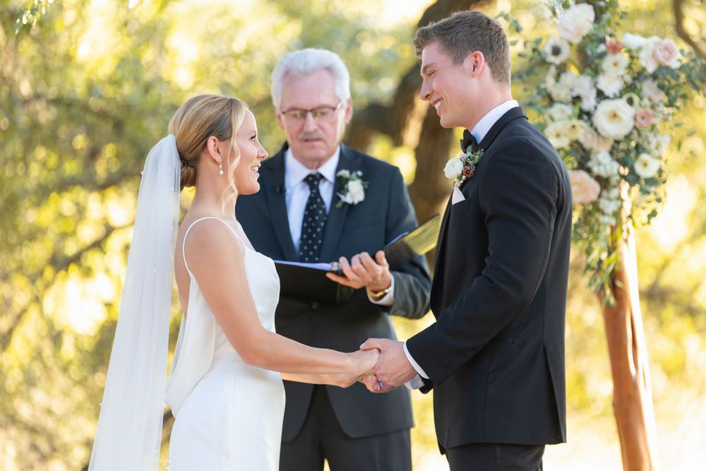 A wedding ceremony takes place outdoors under an elegant floral arch during golden hour.
