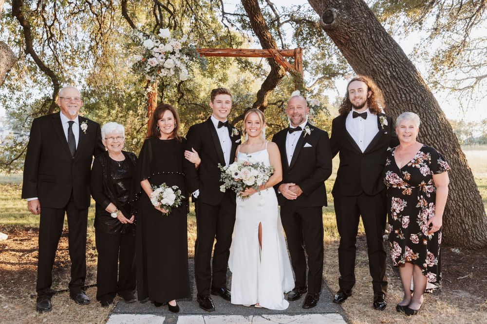 Wedding party poses together outdoors under large tree branches during daytime celebration.