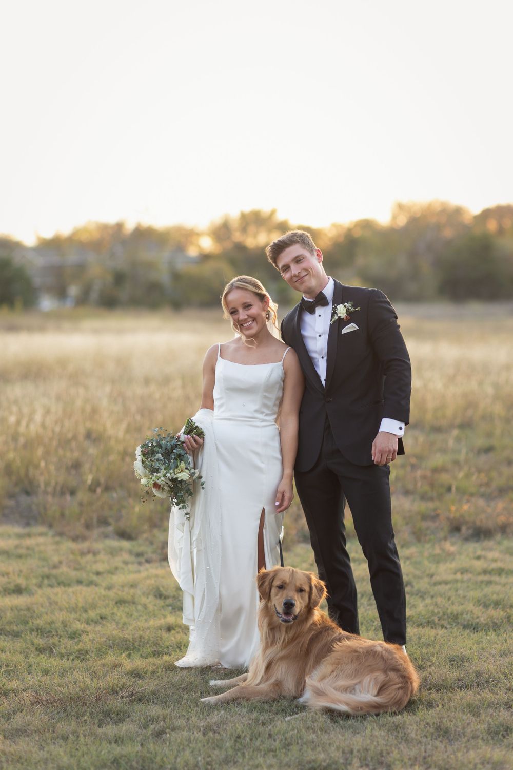 A lovely wedding portrait in a golden field at sunset with a golden retriever sitting at the couple's feet.
