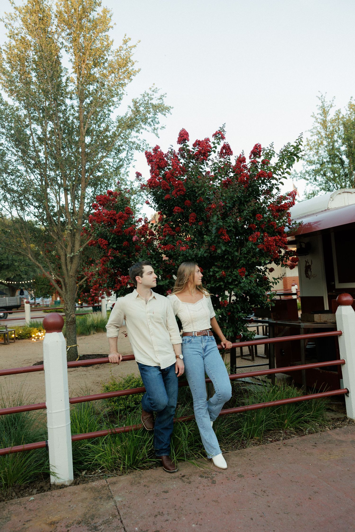 Couple leaning against red fence with blooming crepe myrtle tree in background.