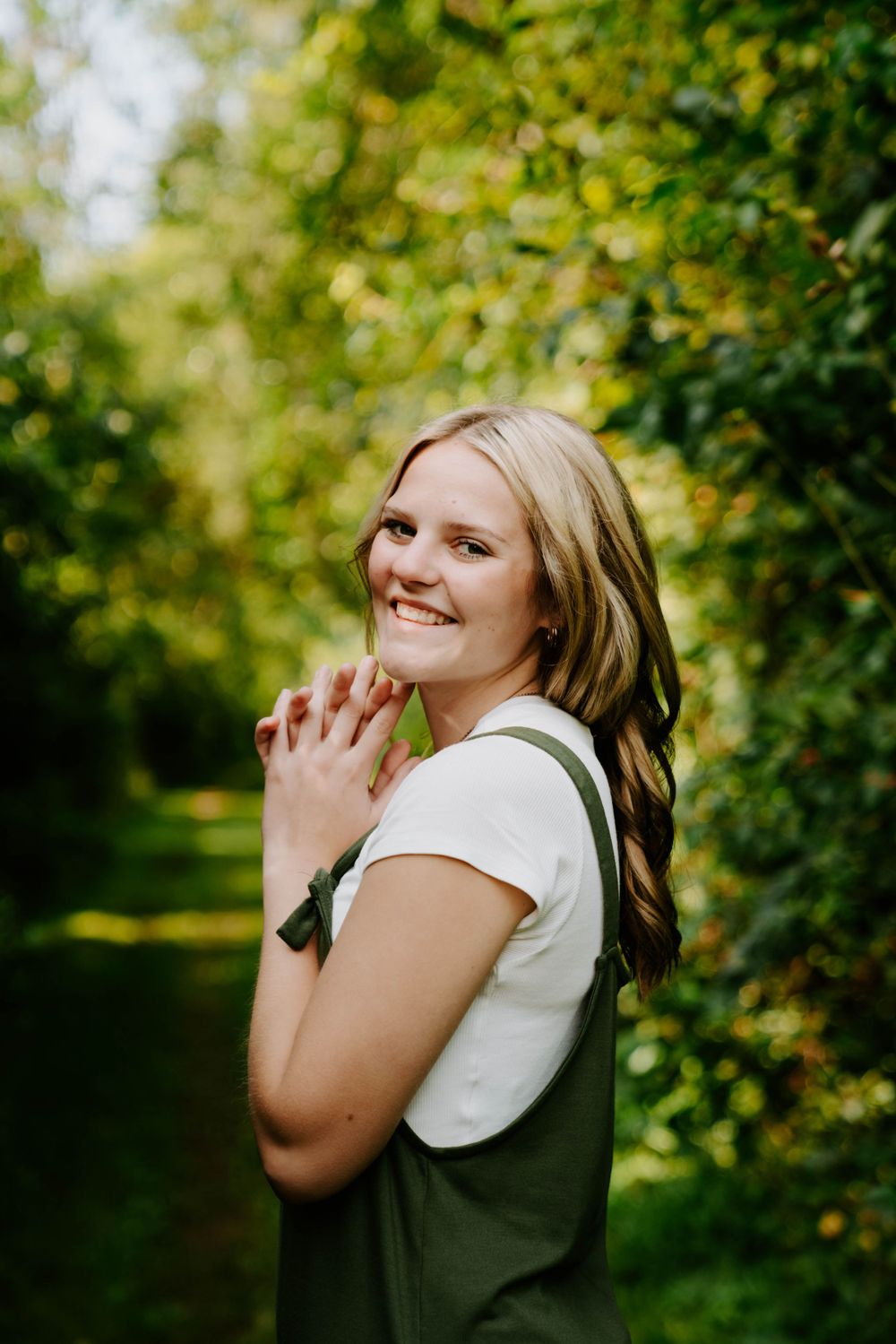 Person in green overalls and white shirt poses joyfully in lush garden setting.