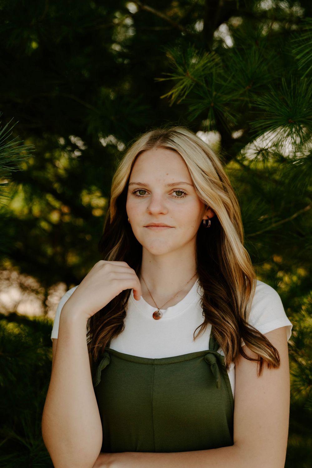 Portrait of person in white shirt and green overalls against natural pine background.