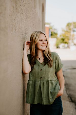 Candid poses against a concrete wall wearing a sage green blouse and dark jeans.