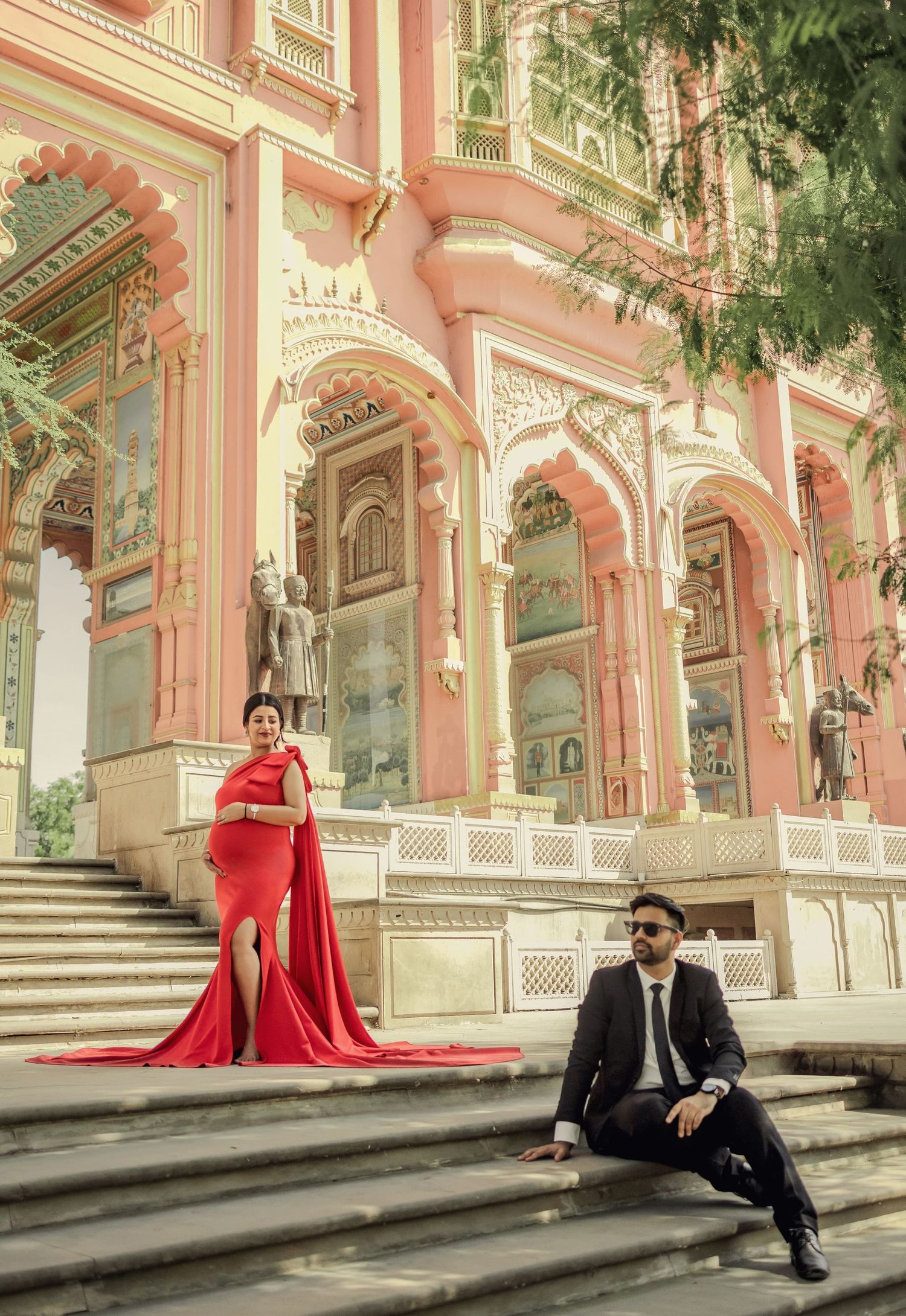 A glamorous photo shoot featuring a red gown against pink palace architecture with detailed arches and columns.