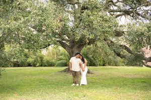 A couple stands together beneath a large, sprawling tree in a serene green park setting.