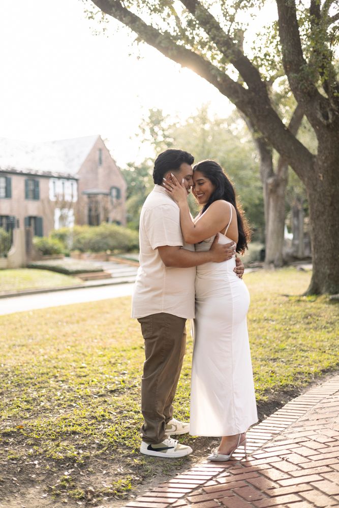 A romantic couple embraces on a brick path surrounded by trees and sunlight during sunset in a residential neighborhood.