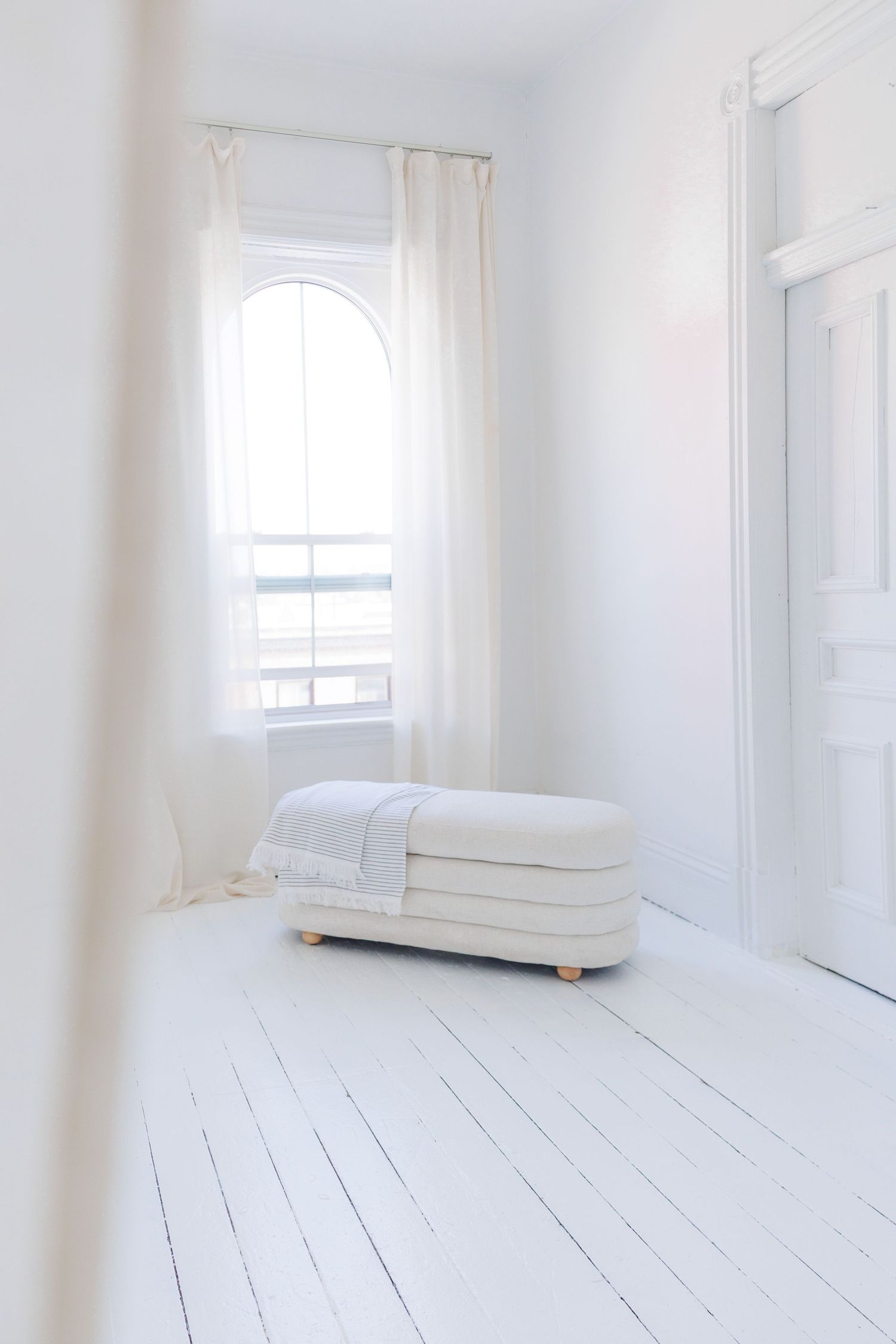 A minimalist white room with two windows, sheer curtains, and a single upholstered armchair in bright natural light.