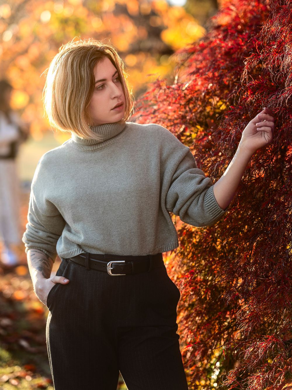 A person in a grey sweater poses against vibrant red autumn foliage during golden hour lighting.