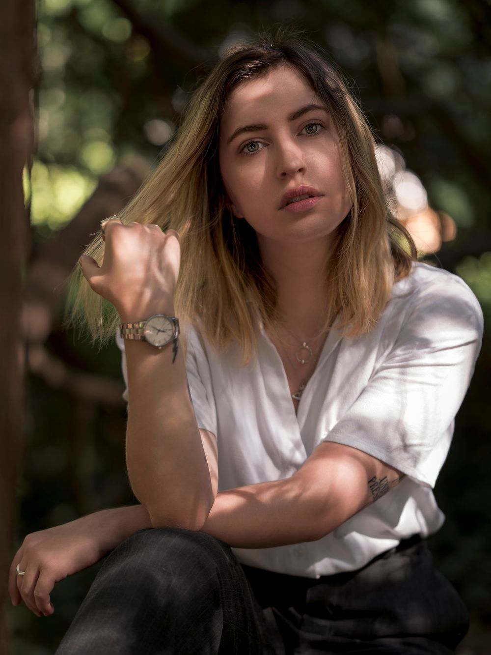 Portrait of someone in a white button-down shirt and dark pants sitting outdoors with natural bokeh background.