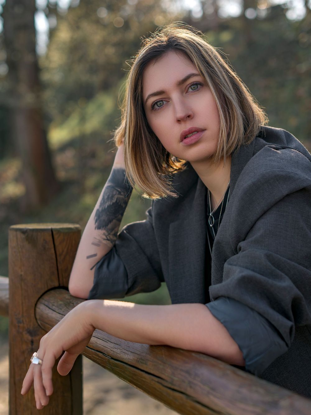 A person in a dark hoodie poses thoughtfully on a wooden railing in an outdoor forest setting.
