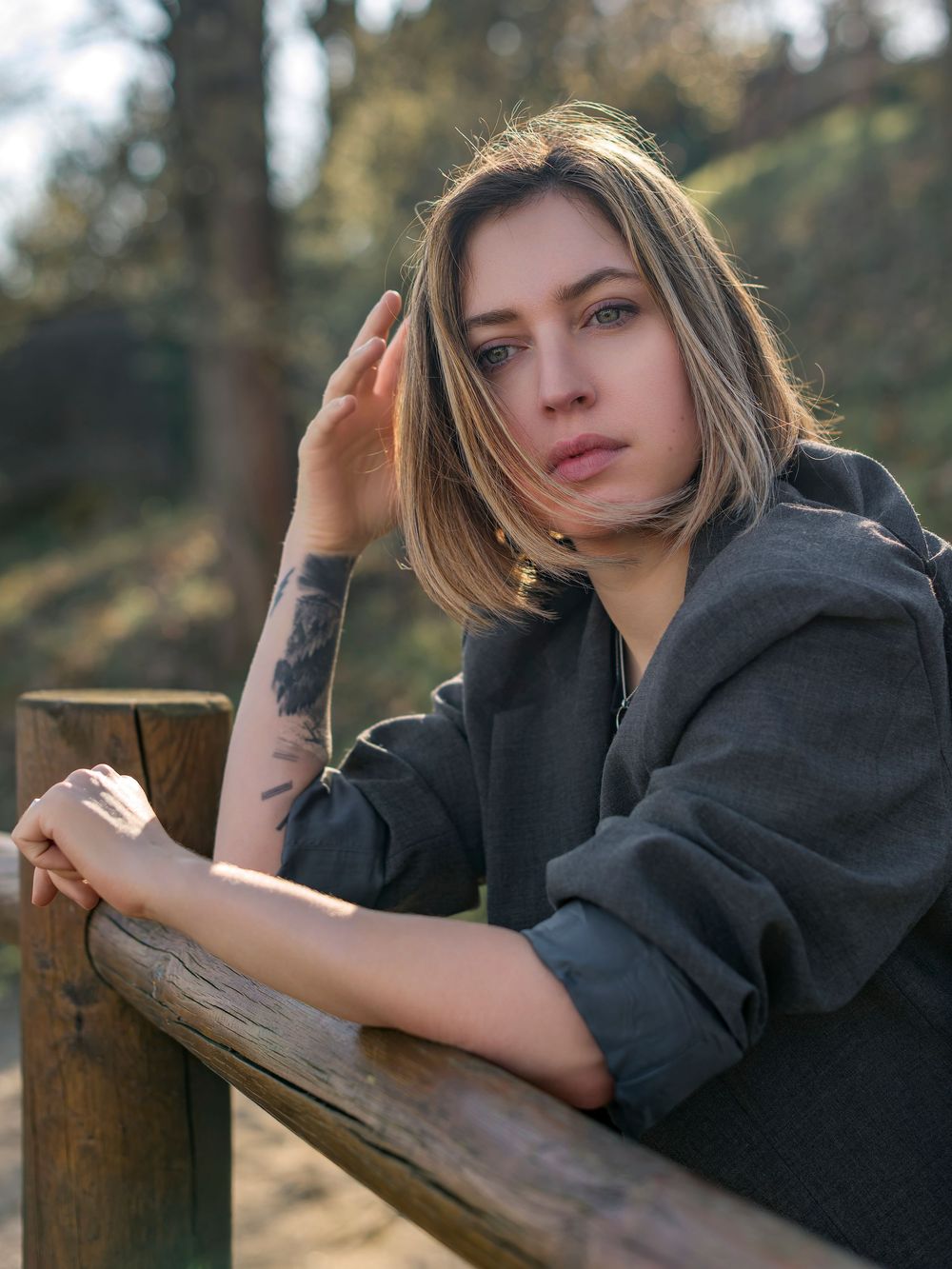 A woman in a black blazer poses thoughtfully against a wooden railing in an outdoor autumn setting.