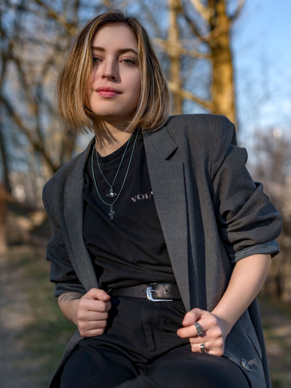 Outdoor portrait showing someone in a gray blazer and black outfit against a blurred natural background in warm lighting.