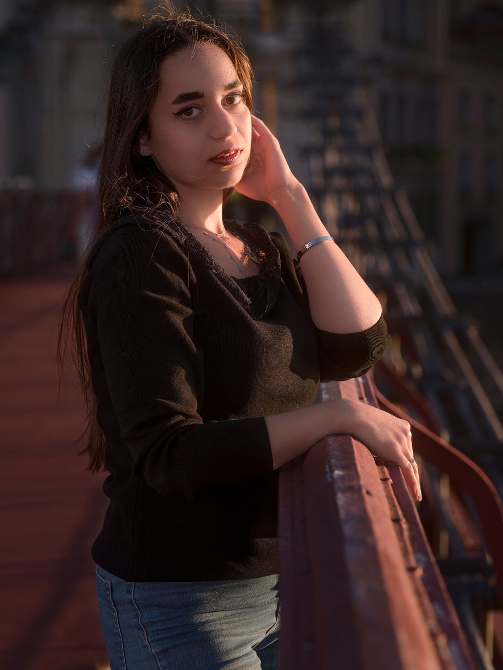 A person in a black sweater and jeans leans against a railing during golden hour sunset lighting.