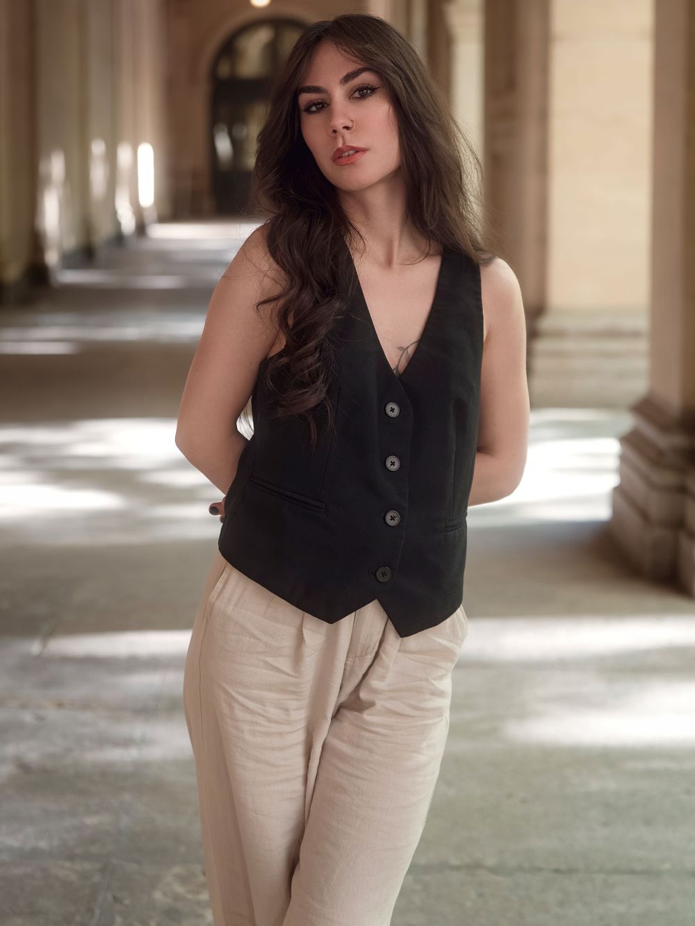 Young woman in black vest and beige trousers posing in elegant archway corridor with stone columns.