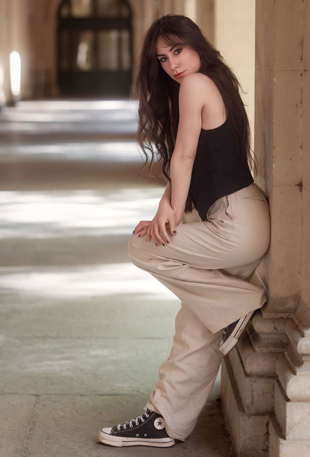 Young woman in black top, beige cargo pants, and Converse sneakers posing gracefully against a stone column.