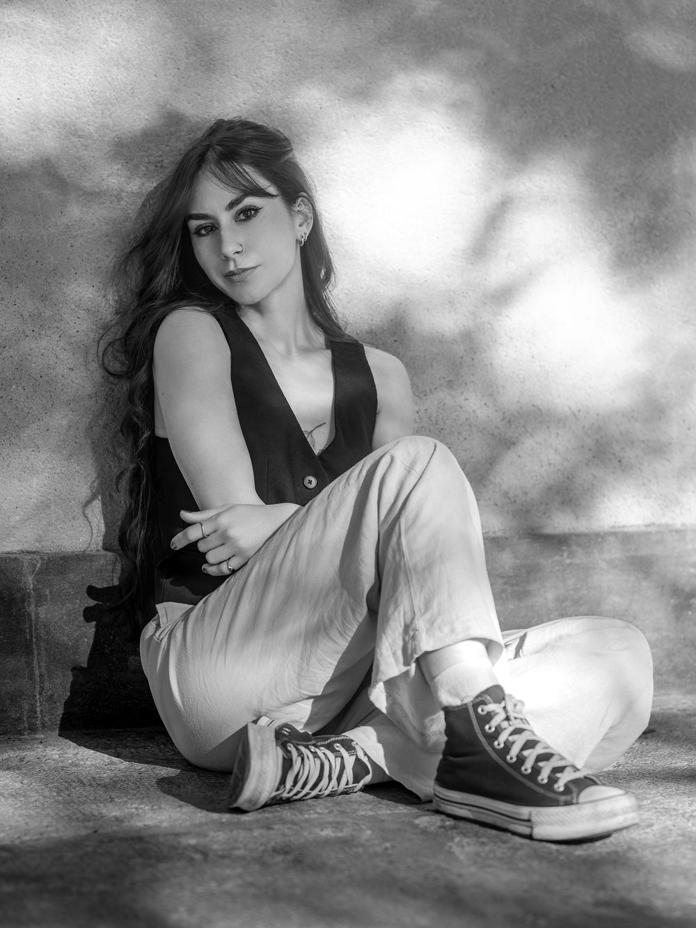 Black and white portrait of a young woman with long hair sitting on the floor wearing casual pants and Converse sneakers.