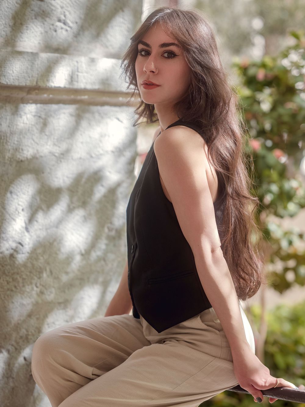 Young woman with long dark hair poses in black sleeveless top and beige pants outdoors near a textured wall.