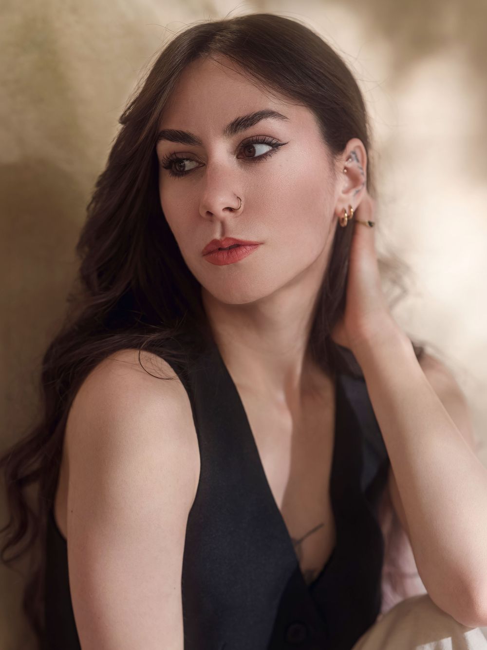 Young woman with dark wavy hair wearing a black dress and gold earrings, posing elegantly against a warm background.
