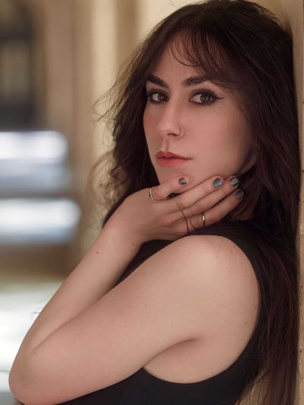 A woman with dark hair leans against a wall, gazing over her shoulder in a black top, rings on her fingers.